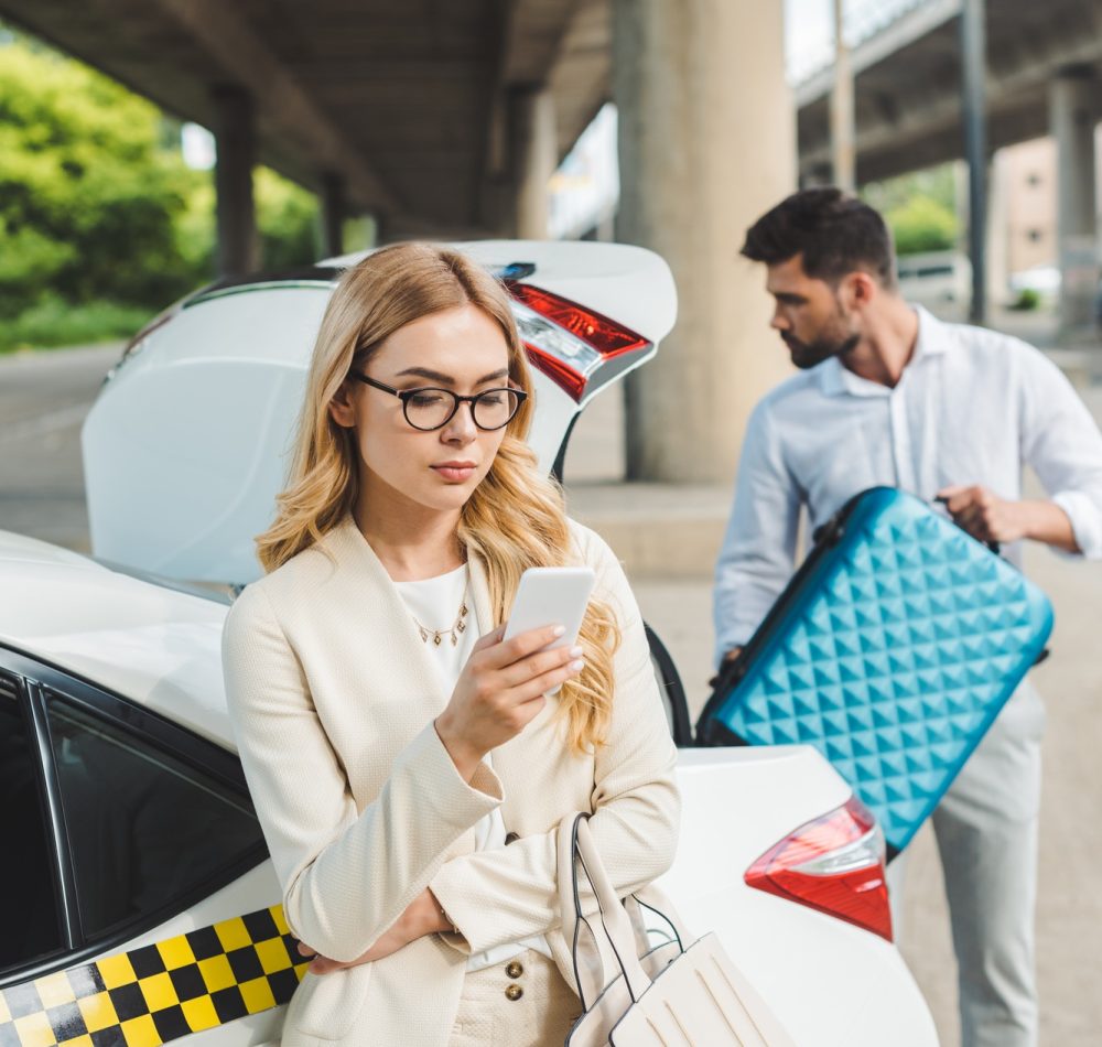 young blonde woman in eyeglasses using smartphone while man putting suitcase in trunk of taxi car